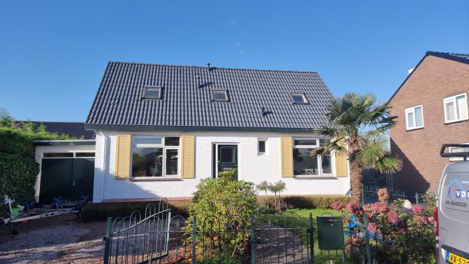 Wit huis met zwarte dakpannen, tuin met bloemen en zonlichte lucht op de achtergrond.